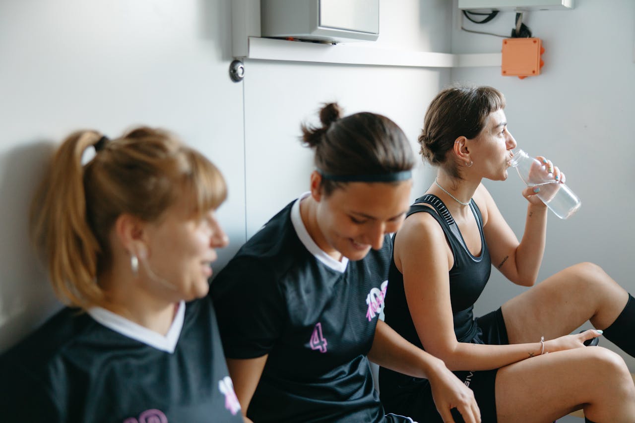 programs-02 Three female athletes in sportswear resting indoors, bonding, and hydrating after a game.